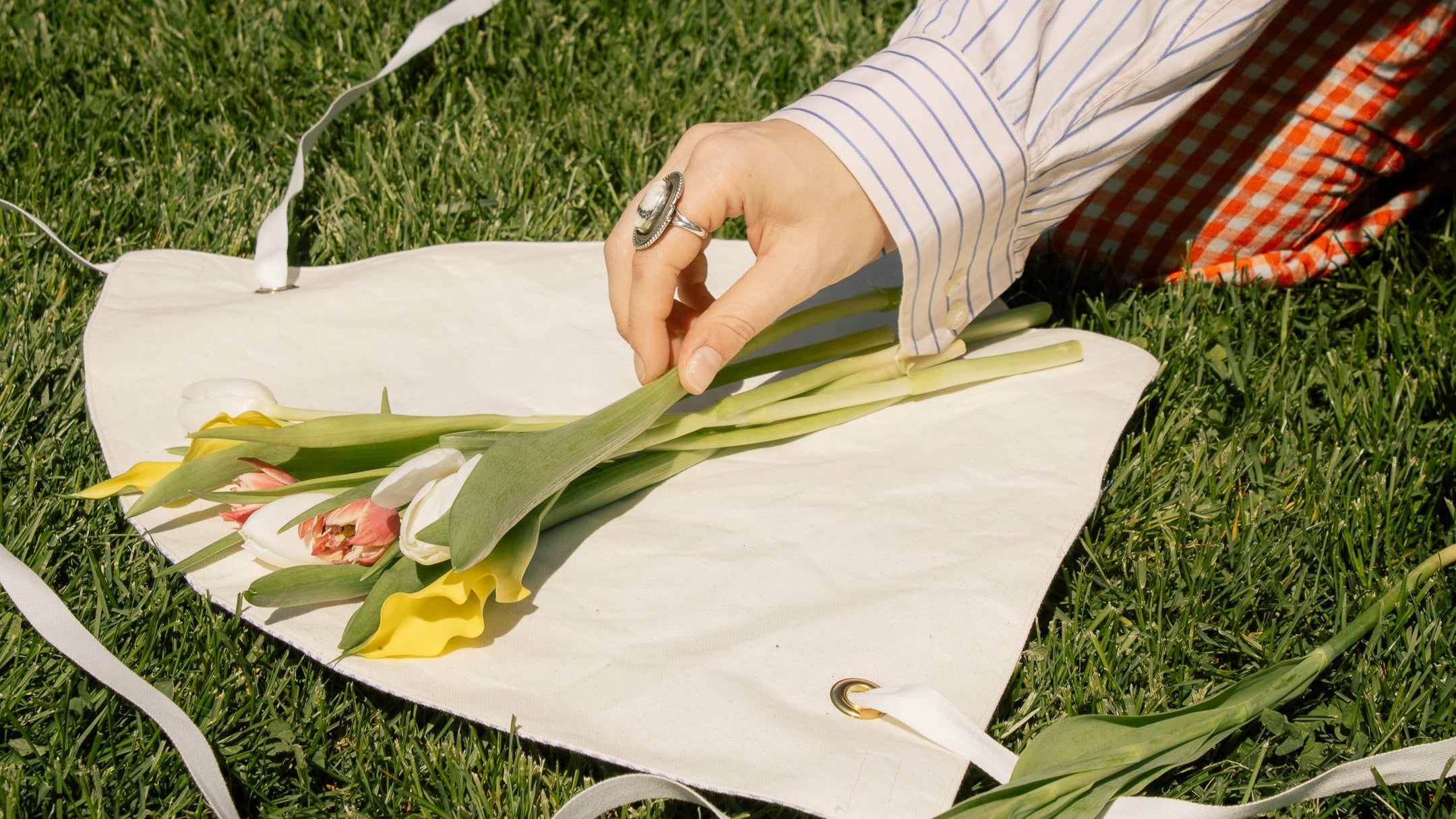 Person arranging pink, white, and yellow tulip flowers on a white canvas bag outdoors on grass